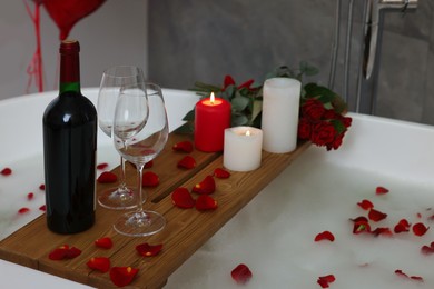 Photo of Wooden tray with wine, burning candles and rose petals on tub in bathroom. Valentine's day celebration