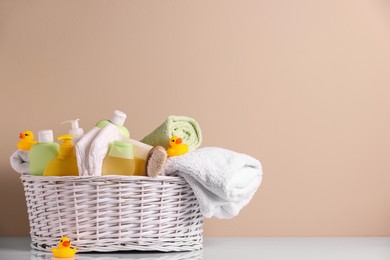 Photo of Wicker basket with baby cosmetic products, bath accessories and rubber ducks on white table against beige background. Space for text