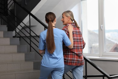 Photo of Young healthcare worker assisting senior woman on stairs indoors, back view
