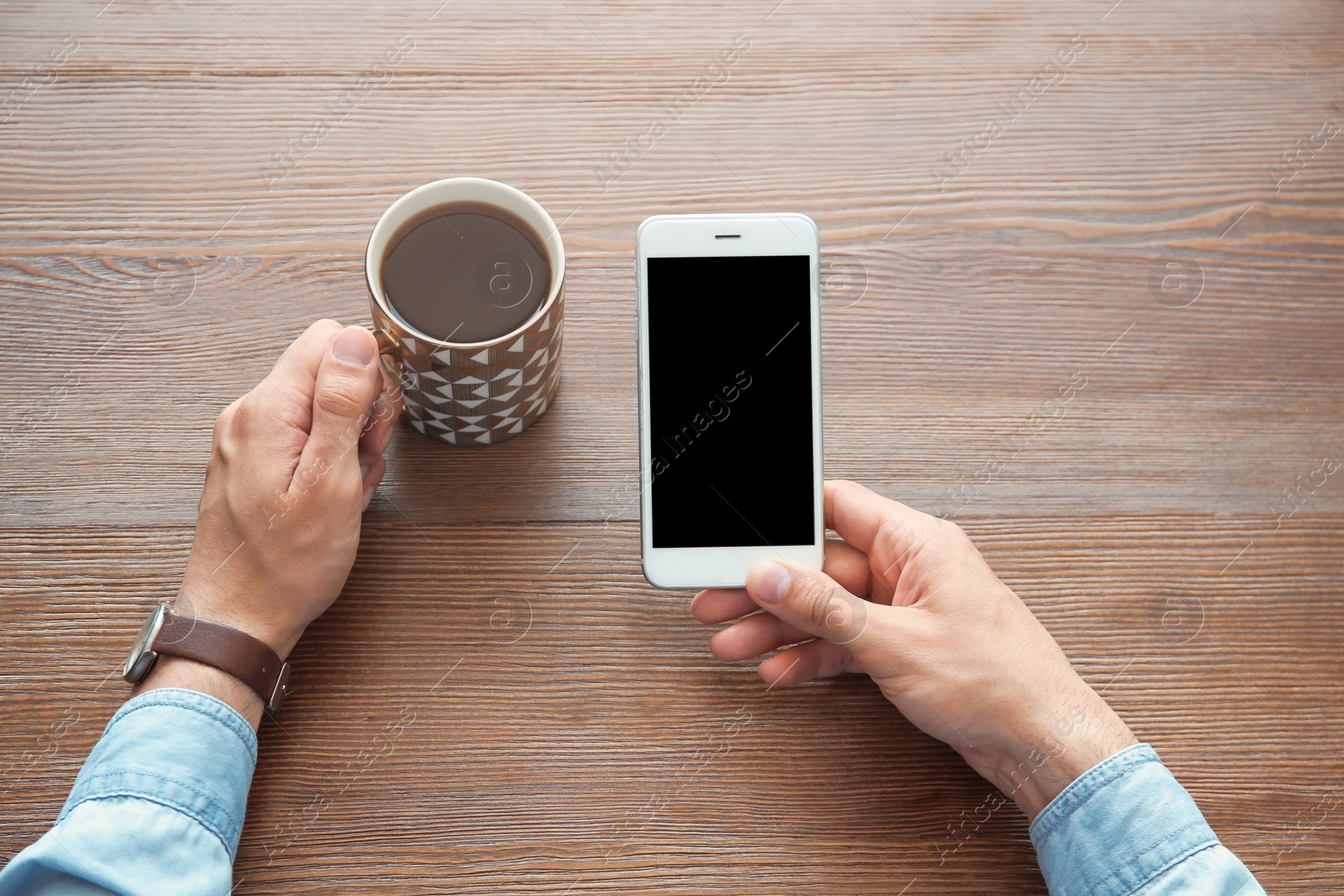 Photo of Young man holding mobile phone with blank screen and cup of coffee in hands at wooden table