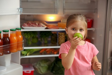 Photo of Cute little girl with green apple near open refrigerator at home