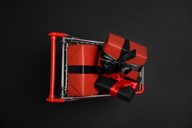Photo of Small shopping cart with wrapped gift boxes on dark background, top view. Black Friday sale