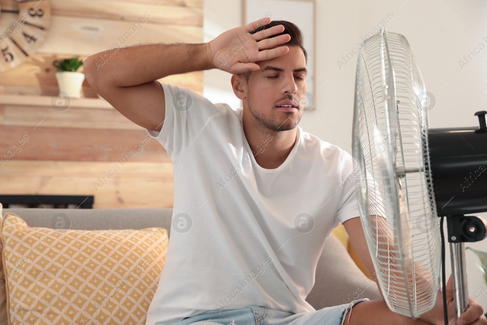 Photo of Man enjoying air flow from fan on sofa in living room. Summer heat