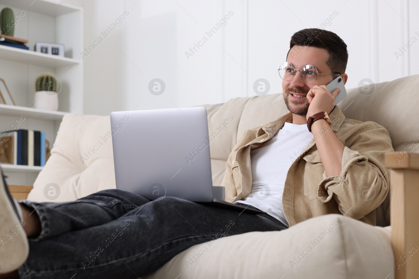 Photo of Happy man talking on phone while working with laptop on sofa at home