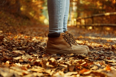 Woman in stylish boots standing on pathway covered with autumn leaves in forest