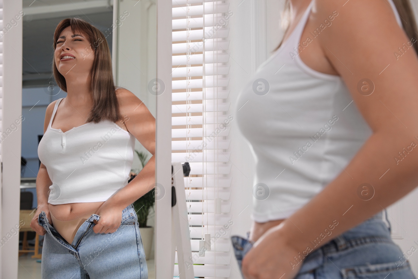 Photo of Young woman struggling to put on tight jeans near mirror at home