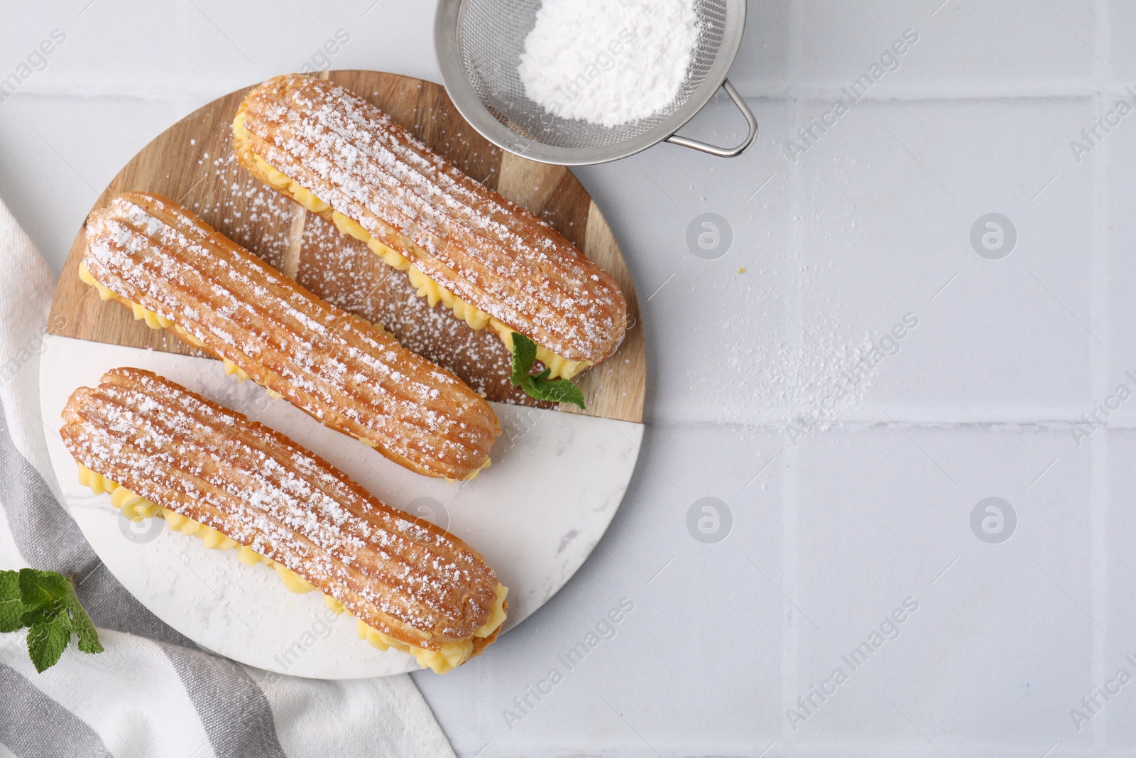 Photo of Delicious eclairs filled with cream and powdered sugar on white tiled table, top view. Space for text