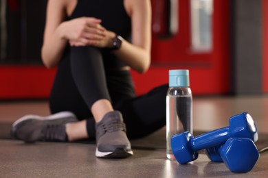 Photo of Young woman sitting on mat in gym, focus on bottle of water and dumbbells