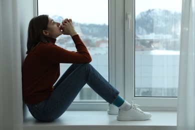 Photo of Sad young woman sitting on windowsill near window at home, space for text