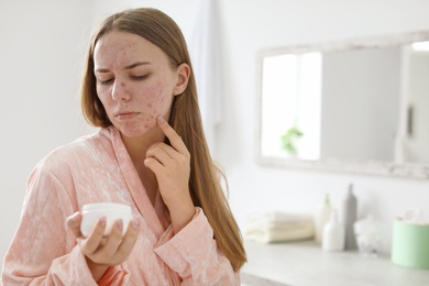 Photo of Young woman with acne problem holding jar with cosmetic product in bathroom