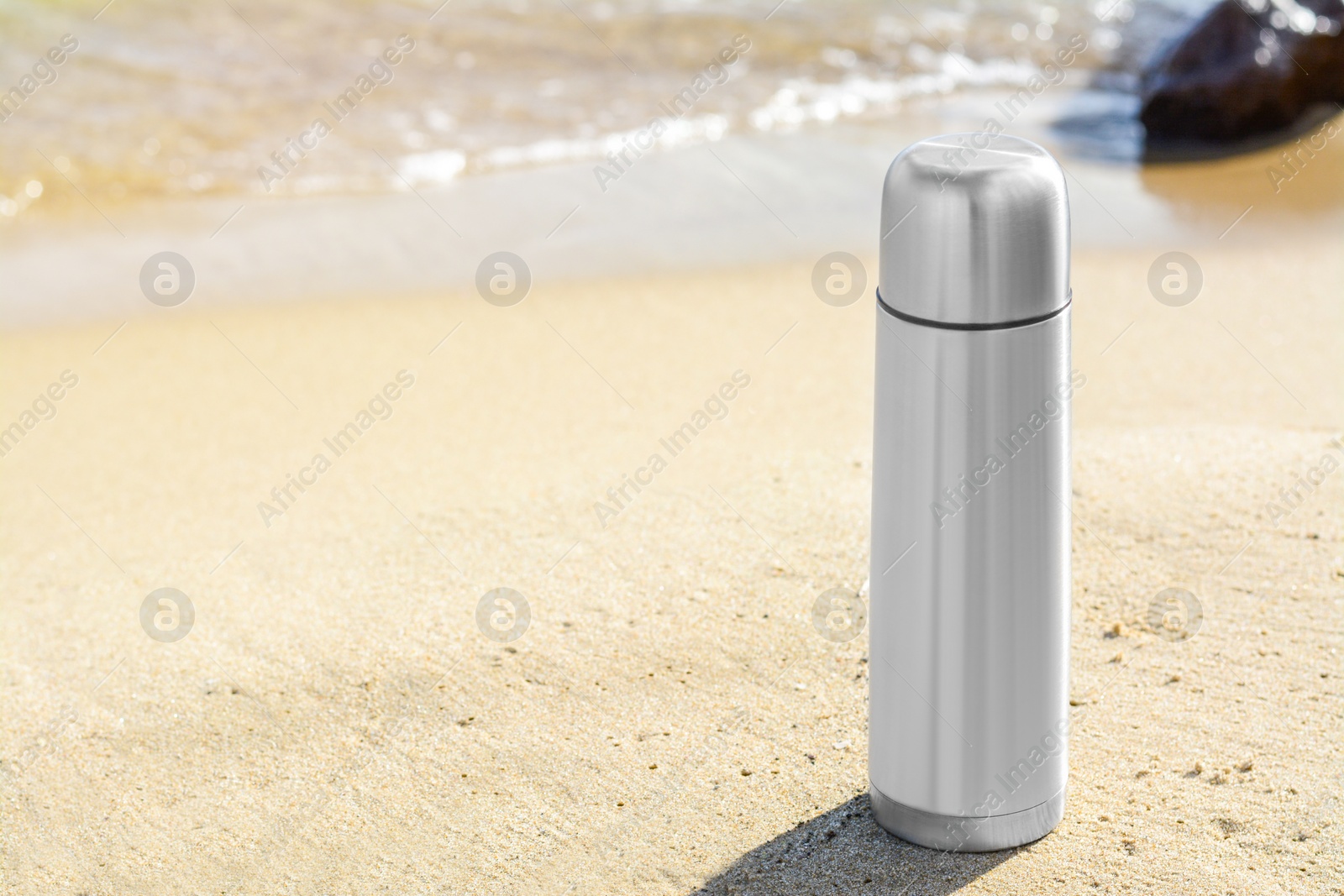 Photo of Metallic thermos with hot drink on sandy beach near sea, space for text