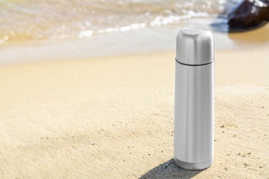 Metallic thermos with hot drink on sandy beach near sea, space for text
