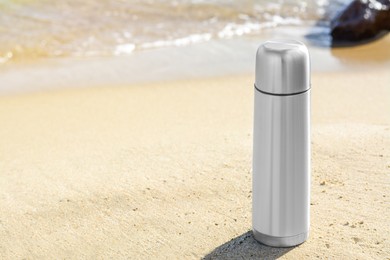 Photo of Metallic thermos with hot drink on sandy beach near sea, space for text