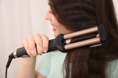 Young woman using modern curling iron against light background, focus on device