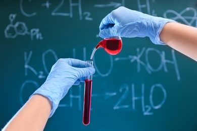 Scientist pouring liquid into test tube against chalkboard, closeup. Chemistry glassware