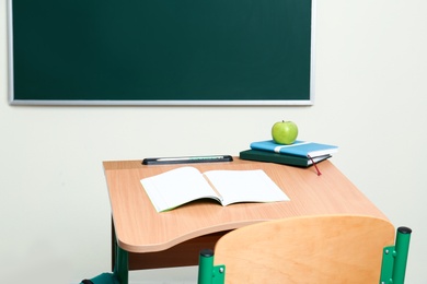 Wooden school desk with stationery, apple and backpack near chalkboard in classroom
