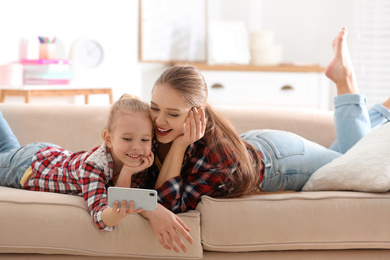 Happy mother and little daughter with mobile phone resting on sofa at home