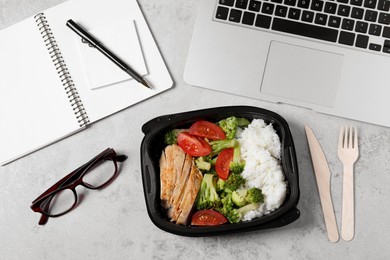 Photo of Container with tasty food, laptop, plastic cutlery and notebook on light grey table, flat lay. Business lunch