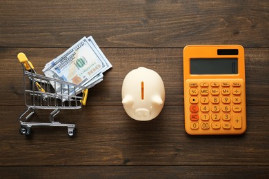 Photo of Calculator, piggy bank and cart with money on wooden table, flat lay