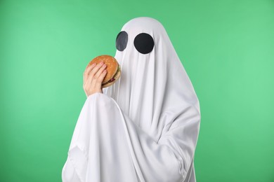 Photo of Creepy ghost. Person in white sheet with tasty hamburger on green background