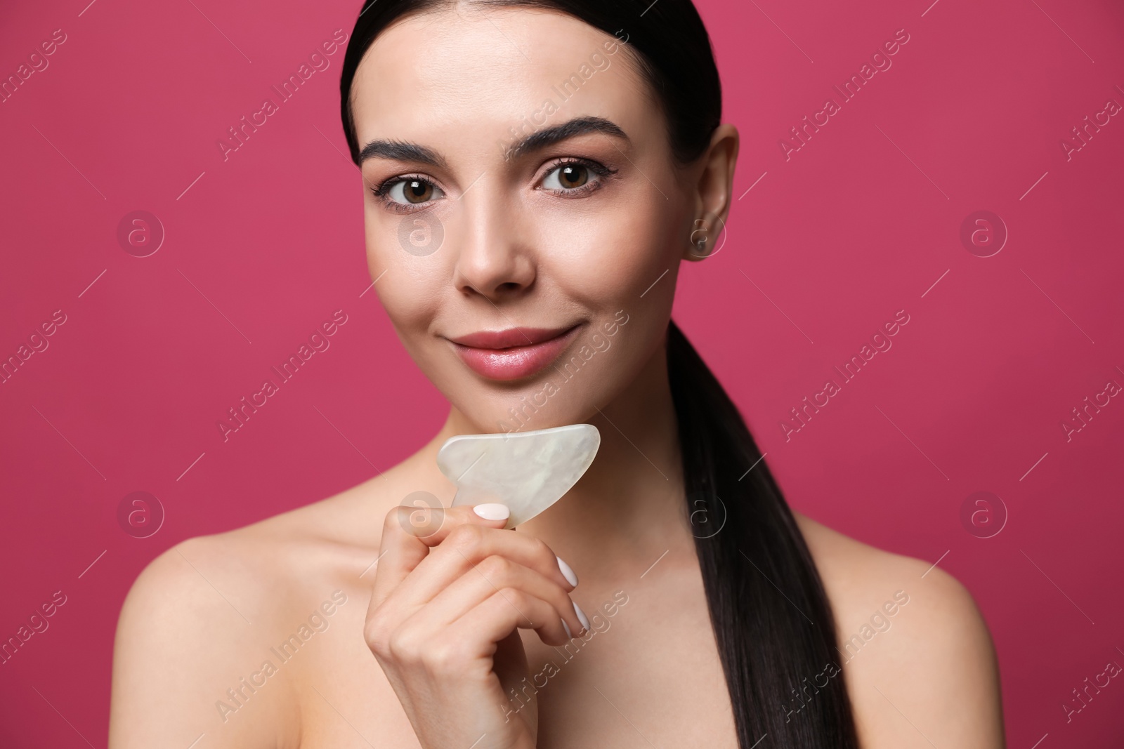 Photo of Beautiful young woman doing facial massage with gua sha tool on pink background, closeup