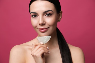 Beautiful young woman doing facial massage with gua sha tool on pink background, closeup