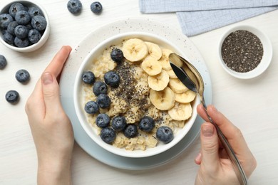 Photo of Woman eating tasty oatmeal with banana, blueberries and chia seeds at white wooden table, top view