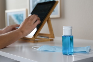 Photo of Hand sanitizer, respiratory mask and blurred woman with tablet on background. Protective essentials during COVID-19 pandemic