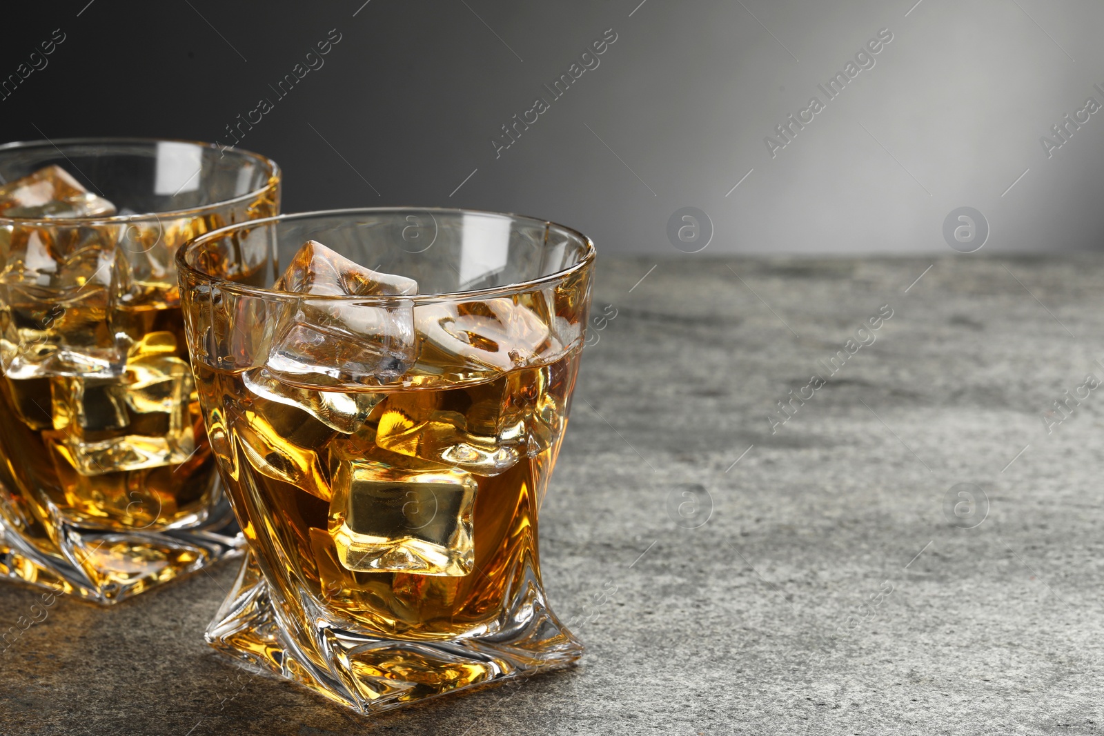 Photo of Whiskey and ice cubes in glasses on grey textured table, closeup. Space for text