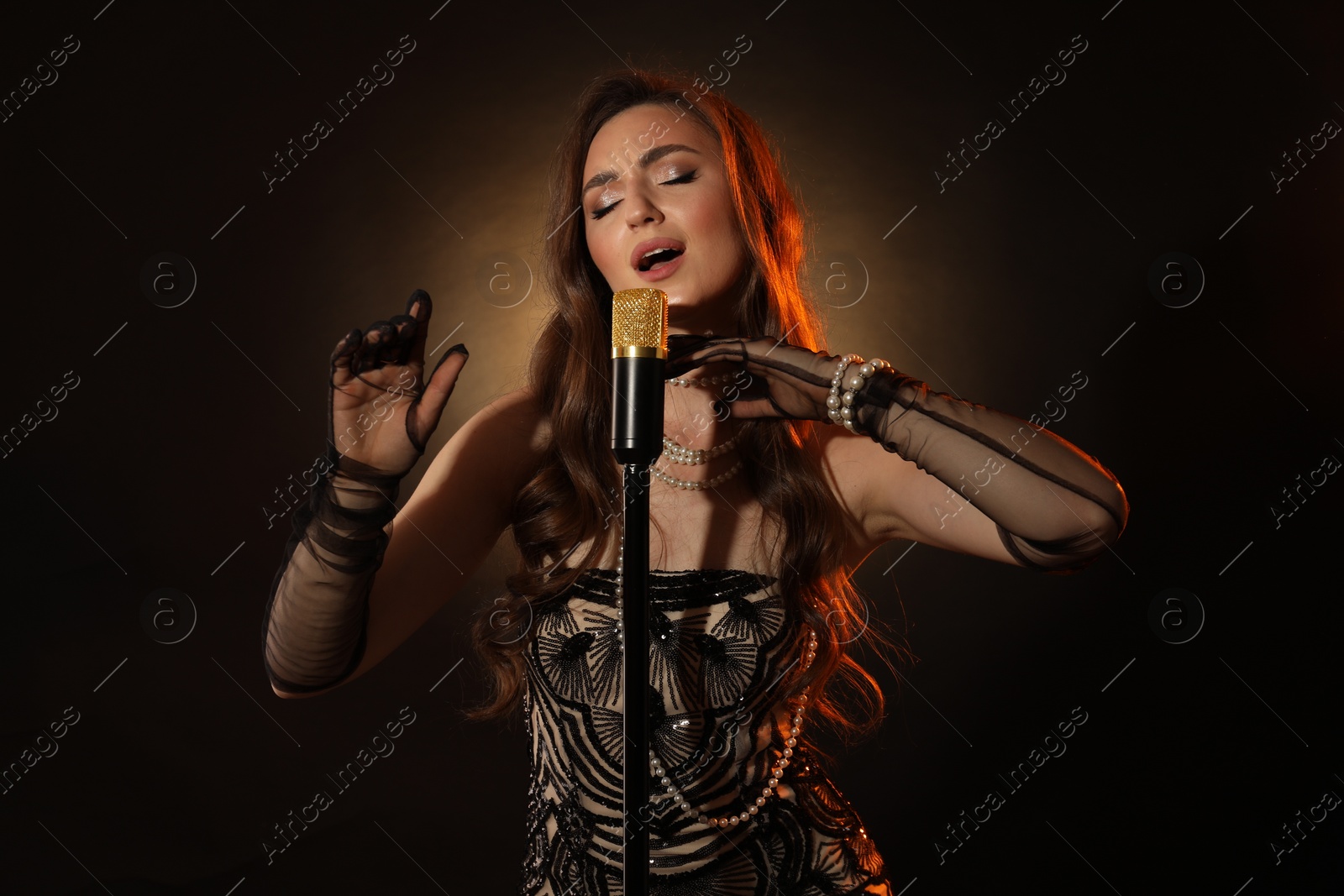 Photo of Beautiful young woman in stylish dress with microphone singing on dark background