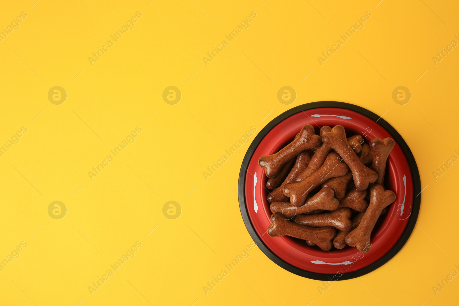 Photo of Red bowl with bone shaped dog cookies on yellow background, top view. Space for text