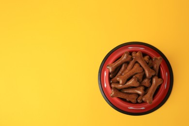 Photo of Red bowl with bone shaped dog cookies on yellow background, top view. Space for text