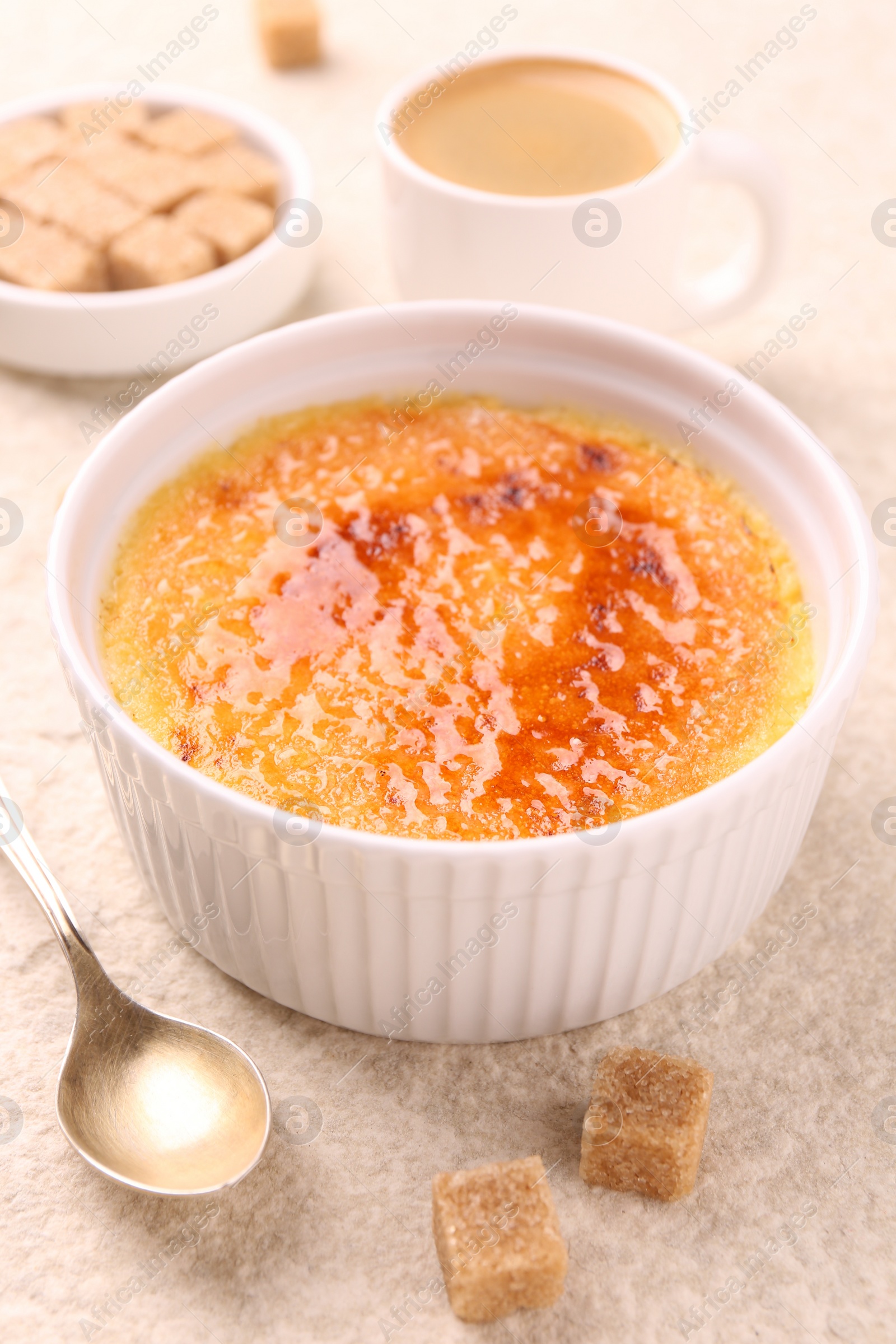 Photo of Delicious creme brulee in bowl, sugar cubes, coffee and spoon on light textured table, closeup