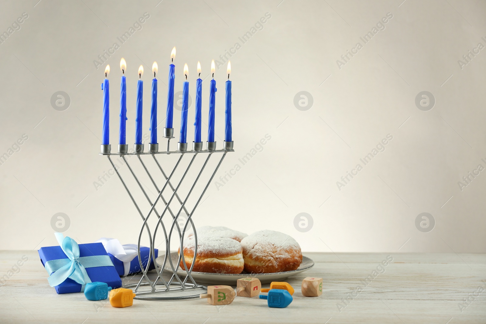 Photo of Hanukkah celebration. Menorah with burning candles, dreidels, donuts and gift boxes on wooden table against light grey background, space for text