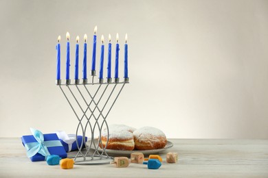 Hanukkah celebration. Menorah with burning candles, dreidels, donuts and gift boxes on wooden table against light grey background, space for text