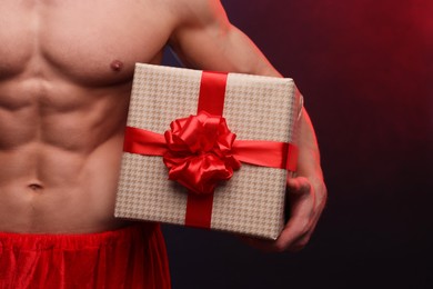 Young man with muscular body holding Christmas gift box on color background, closeup