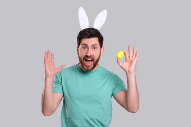 Emotional man in cute bunny ears headband with Easter egg on light grey background