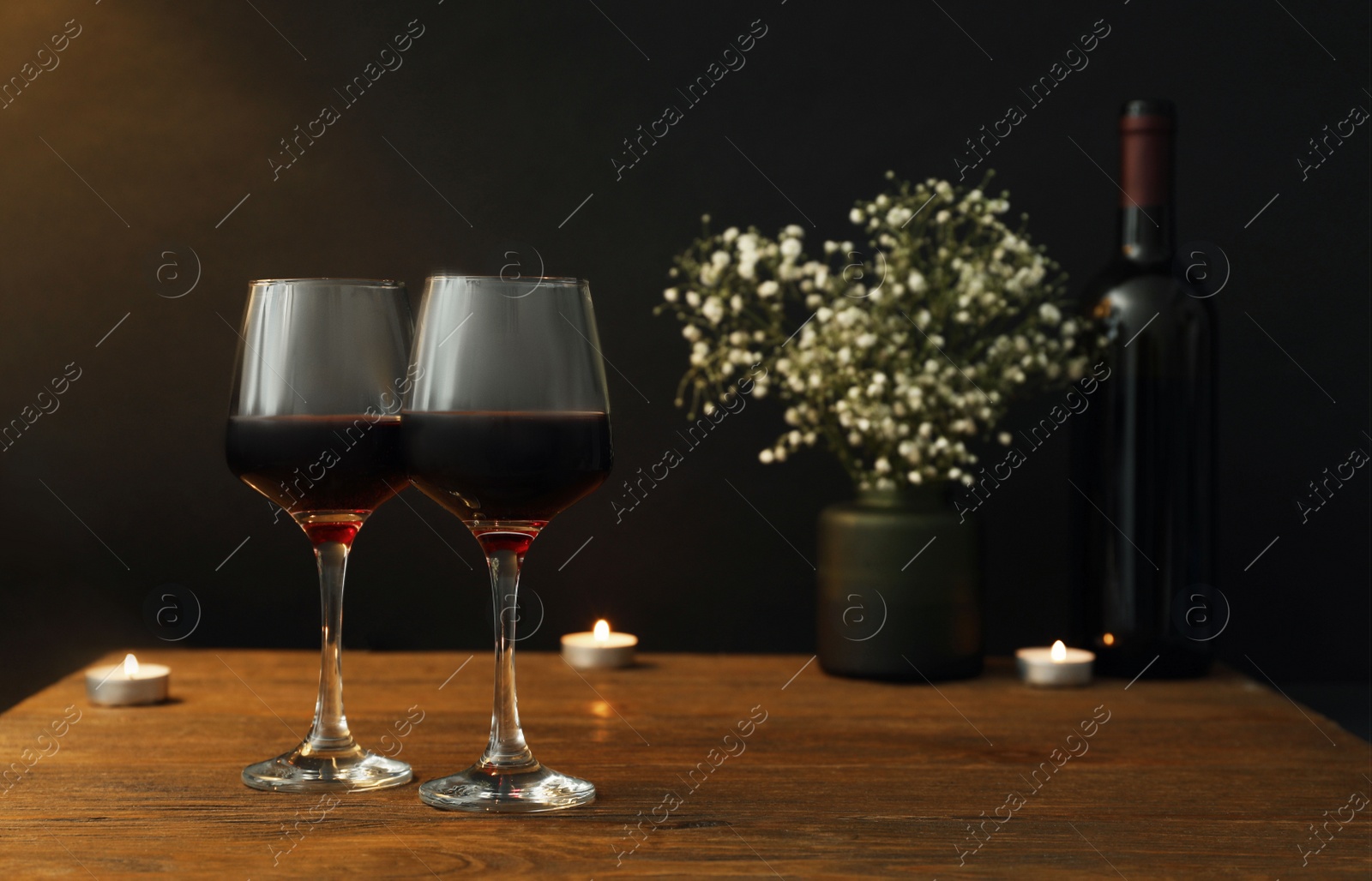 Photo of Glasses of wine, candles and flowers on wooden table near dark wall