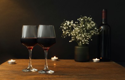 Photo of Glasses of wine, candles and flowers on wooden table near dark wall