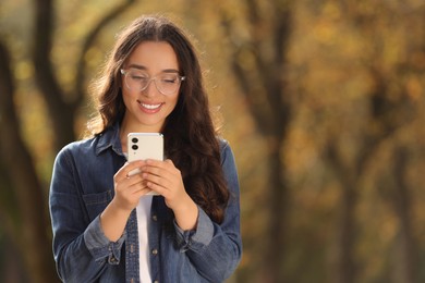 Young woman using smartphone in autumn park, space for text