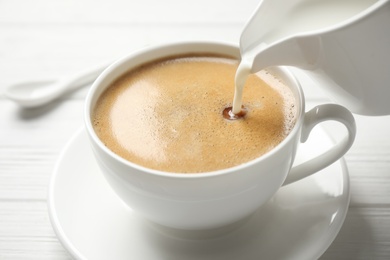 Photo of Pouring milk into cup of hot coffee on white wooden table, closeup