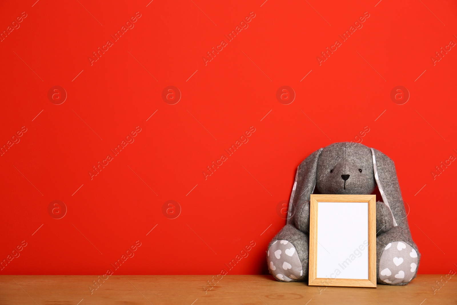 Photo of Empty photo frame and and soft rabbit on wooden table against red background, space for text. Child room interior