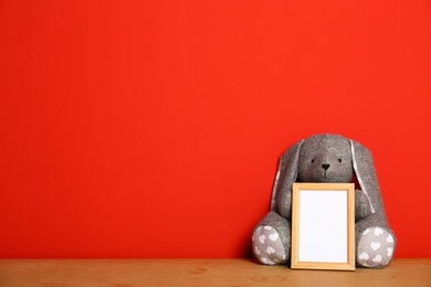 Photo of Empty photo frame and and soft rabbit on wooden table against red background, space for text. Child room interior