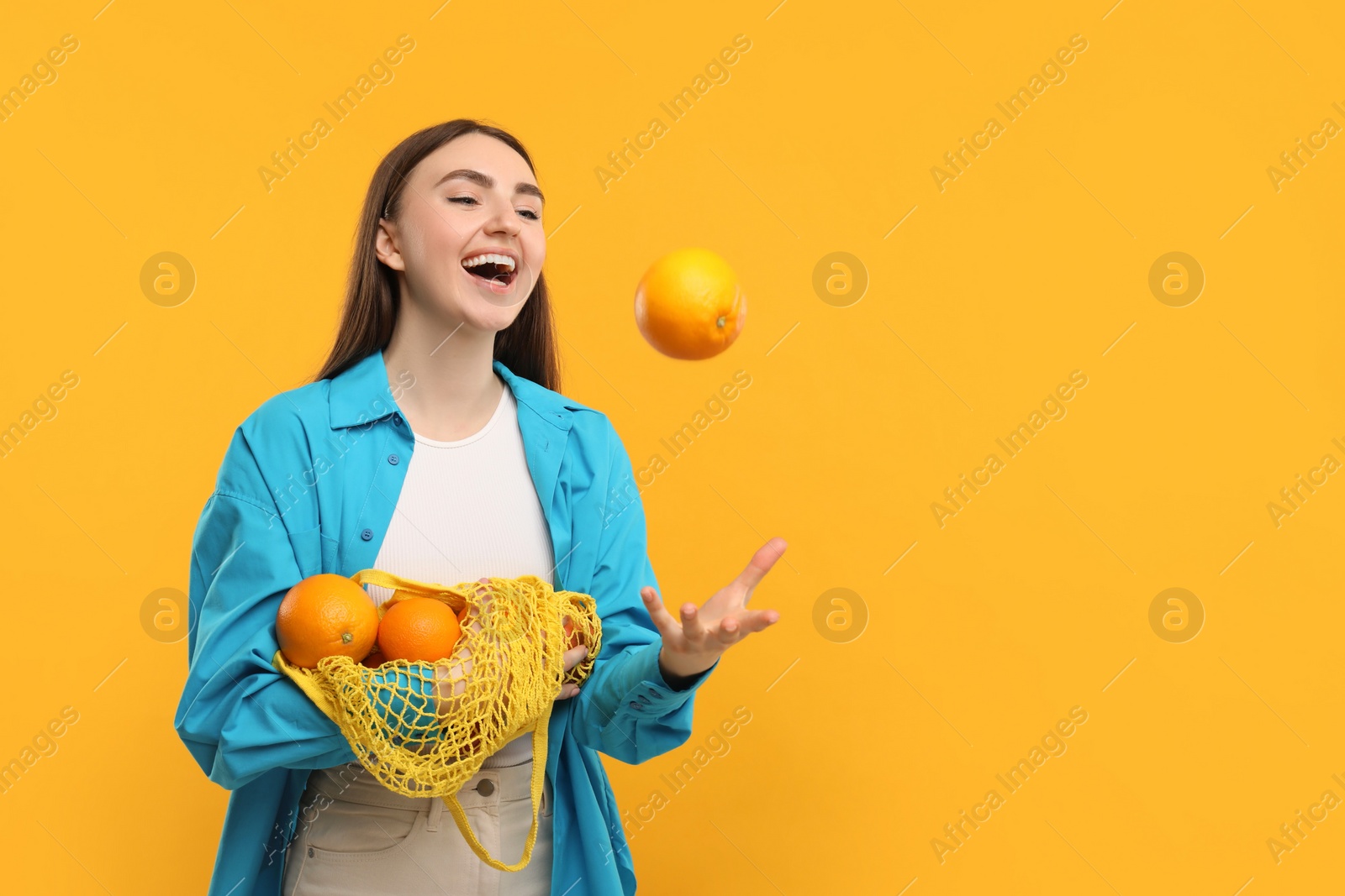 Photo of Woman with string bag of fresh oranges throwing fruit on orange background, space for text