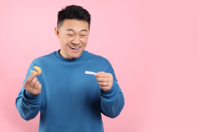 Emotional asian man holding tasty fortune cookie and reading prediction on pink background. Space for text