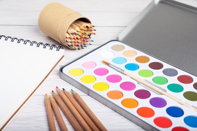 Watercolor palette with brush, colorful pencils and notebook on white wooden table, closeup