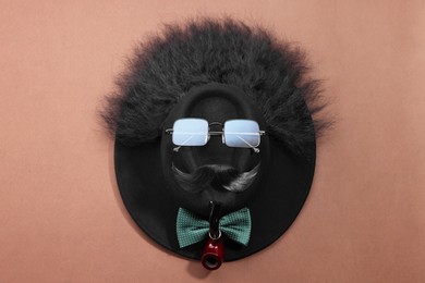 Photo of Man's face made of artificial mustache, sunglasses and hat on brown background, top view