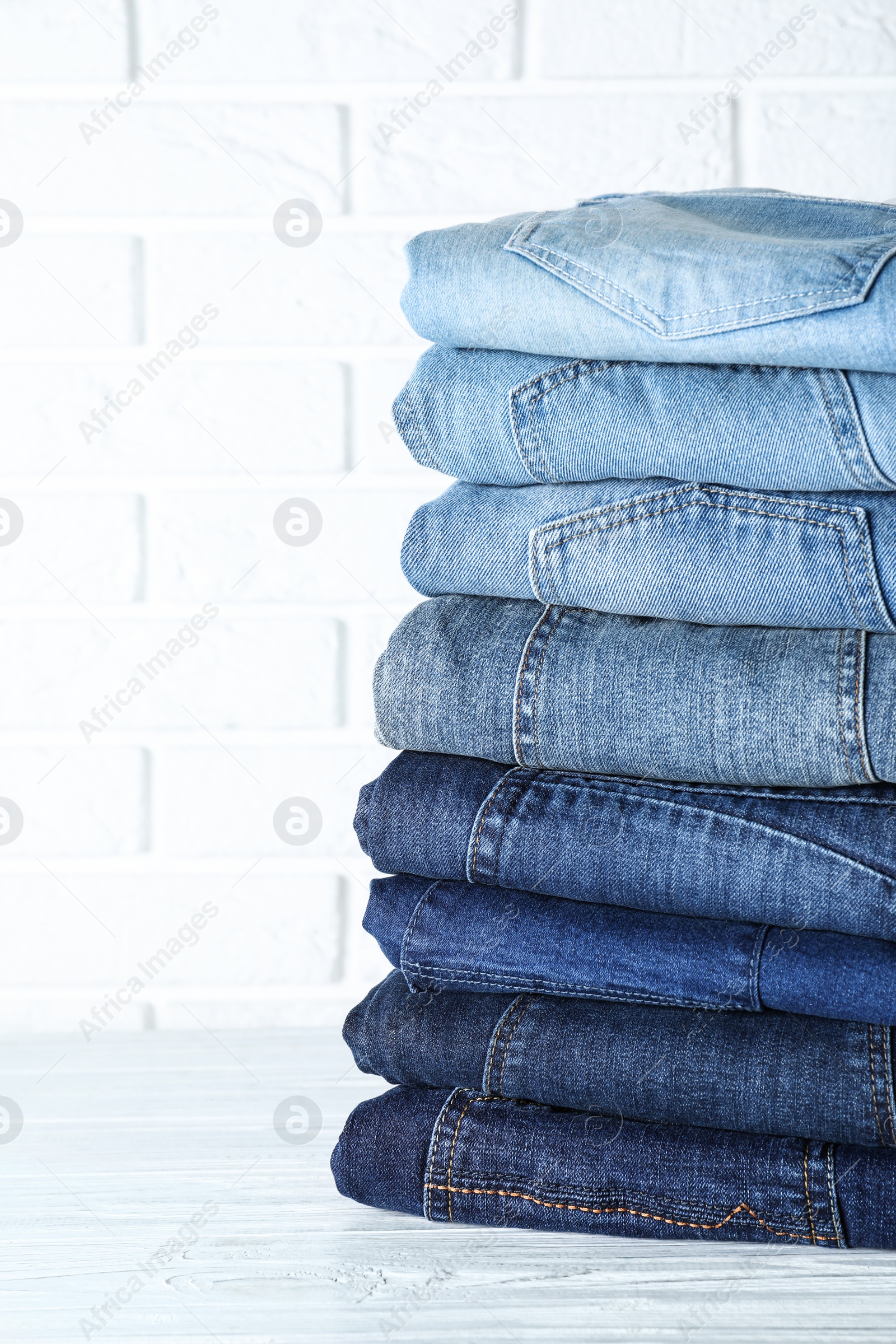 Photo of Stack of different jeans on white wooden table against brick wall