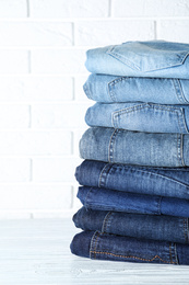 Photo of Stack of different jeans on white wooden table against brick wall