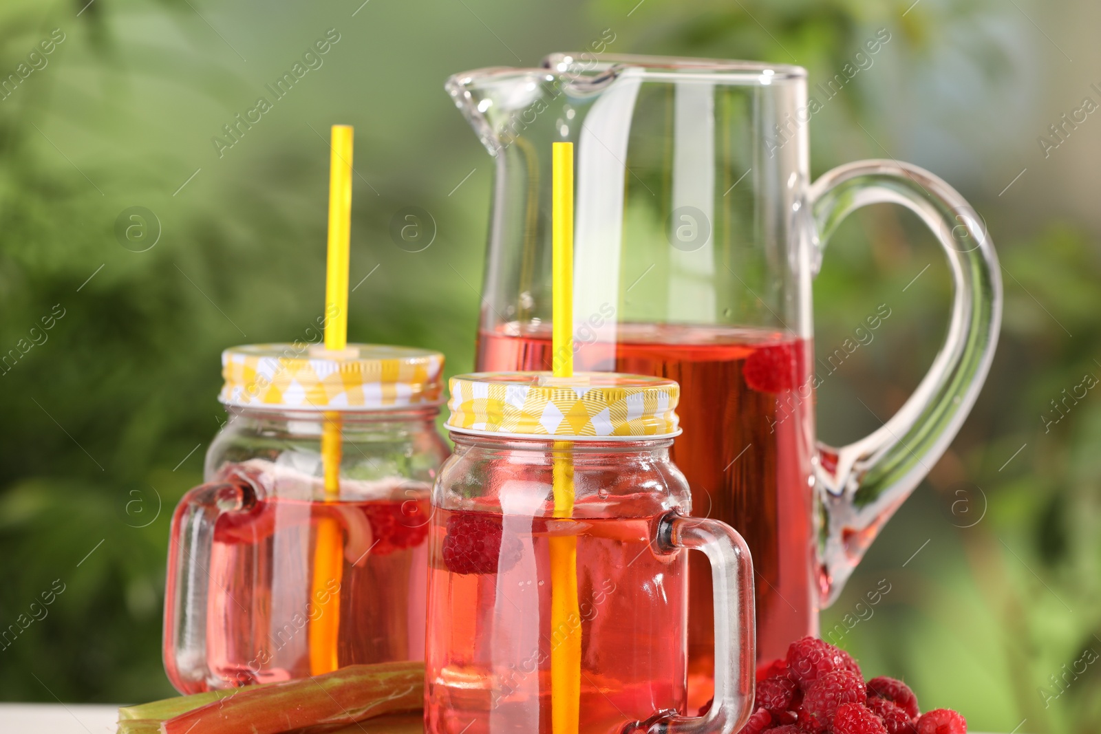 Photo of Mason jars and jug of tasty rhubarb cocktail with raspberry outdoors, closeup